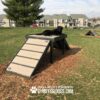 A black dog lies on top of an outdoor agility ramp in a grassy area with apartment buildings in the background. The text "Gyms for Dogs" is displayed at the bottom of the image.