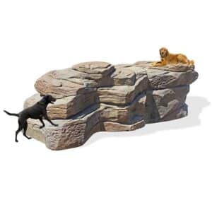 Two dogs, one black and one golden, play on a large artificial rock structure. The black dog climbs up from the left, while the golden dog lies on top of the rocks with a yellow toy.