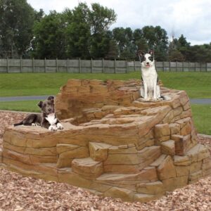 Two dogs rest on a large artificial rock structure in a park, surrounded by wood chips and green trees in the background. One dog is lying down while the other sits alertly on a higher ledge.