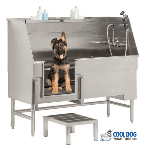 A German Shepherd puppy sits in the Dog Wash Tub Pro Series Parallel Access Tub with grooming products on a shelf and a handheld showerhead attached, while a small step stool is placed in front of the tub.