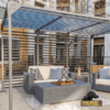 A modern patio with a gray woven sofa, matching armchair, and coffee table sits beneath The Dog Park Shade - 8' x 8' metal pergola. Decorative pillows, a lantern, and a black dog add coziness. The "Dog Park Outfitters" logo is visible.