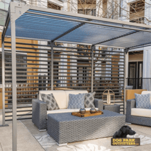 A modern patio with a gray woven sofa, matching armchair, and coffee table sits beneath The Dog Park Shade - 8' x 8' metal pergola. Decorative pillows, a lantern, and a black dog add coziness. The "Dog Park Outfitters" logo is visible.