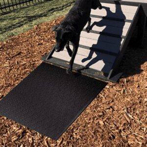 A black dog climbs down a small ramp with stairs onto a Jump Dog Agility Mat, set outdoors on wood chips with grass visible in the background.