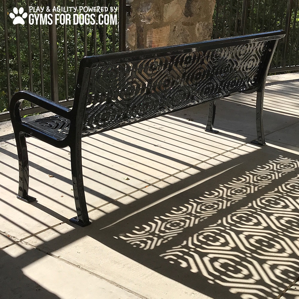 A decorative 4ft Luxury Park Bench made of thermoplastic-coated steel casts intricate geometric shadows in the sunlight; a railing and greenery are in the background. “Gyms for Dogs” appears in the top left corner of the image.