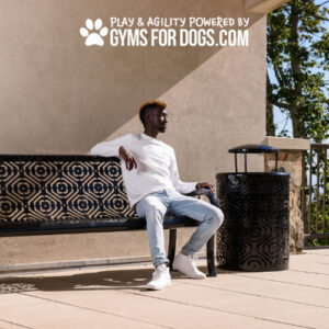 A man in a white shirt and light jeans sits on a patterned outdoor bench near a black trash can, with sunlight casting shadows. "Play & Agility Powered by Gyms For Dogs.com" is displayed above him.