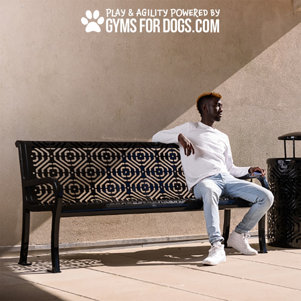 A man in a white shirt and jeans sits on The Luxury Park Bench - Thermoplastic Coated Steel - 6ft near a wall. Above him, text reads, "Play & Agility Powered by Gyms for Dogs.com" with a paw print icon.