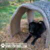 A black dog is lying on wood chips under a curved concrete tunnel in a fenced outdoor area. Text reads: "Play & agility powered by GYMSFORDOGS.COM" with a paw print icon.