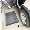 A black mat with a blue Jump Dog logo is placed on the ground next to the open rear door of a white car in a garage. The Jump Dog website and logo appear in the corner of the image.