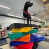 A black dog wearing a red bandana stands confidently on top of a tall stack of colorful gym mats in a spacious, modern indoor room with office furniture and equipment.