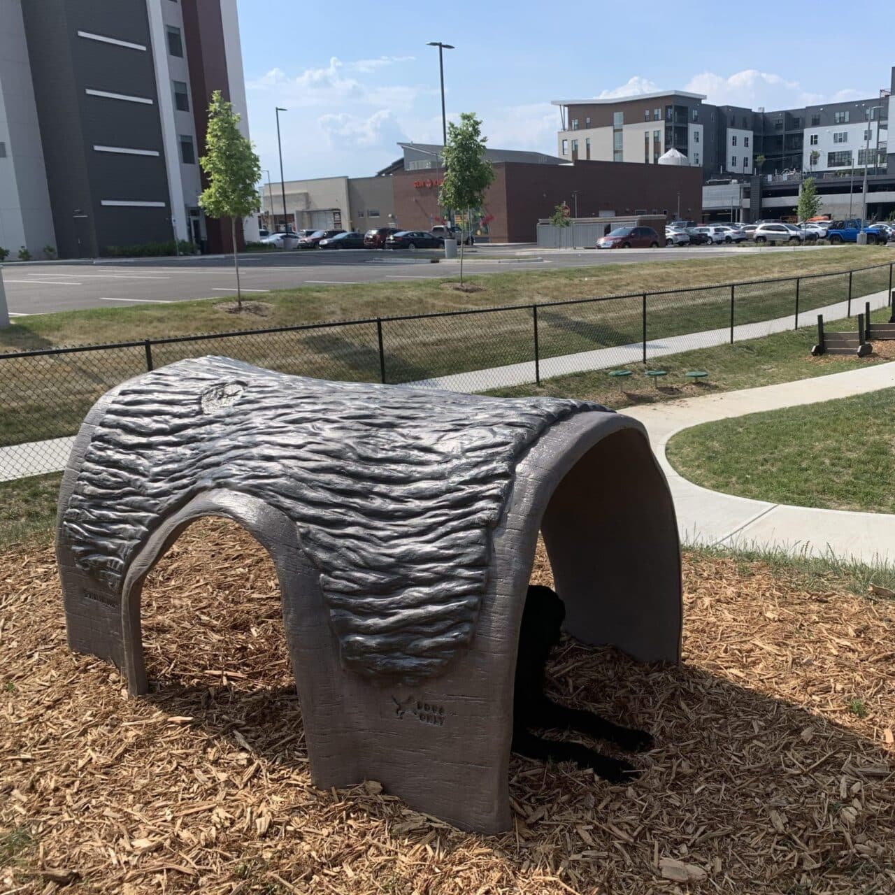 A large, gray, cave-shaped structure sits on wood chips in a park near a sidewalk. A black dog lies in its shade, partially visible. Buildings, trees, parked cars, and a fence are in the background under a sunny sky.