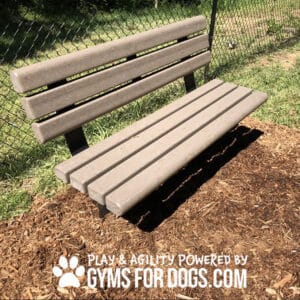 A Trail Bench w/ Back - In-Ground Mount made of tan wooden slats sits on mulch near a chain-link fence in a grassy area. Text below reads: "Play & Agility Powered by GymsForDogs.com" with a paw print symbol.