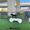 A black dog stands atop The Agility Bone - Free Standing Heavy- Antique Polar (L/X), a large white bone-shaped structure labeled "Gyms For Dogs," in a fenced outdoor area with artificial grass beneath an overpass.