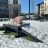 A dog in a pink sweater stands on the Bridge Climb 24" Tall (L/X) ramp at the dog park, surrounded by snow and modern apartment buildings in the background.