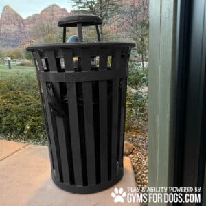 A black Luxury Park Waste Receptacle - Bonnet Lid stands on a sidewalk near greenery and red rock mountains. The bottom corner features the "Play & Agility Powered by Gyms for Dogs" logo with a paw print.