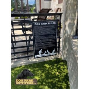 A Dog Park Rules Sign - 36" W x 42" H hangs on a black metal fence next to outdoor seating, with green artificial turf underfoot. A logo featuring a black dog and "Dog Park Outfitters" is in the bottom left corner.