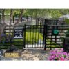 A gated entrance to a dog park with a "Welcome to the Dog Park" sign on the fence. Green grass, trees, and flowers are visible inside. A "Dog Park Outfitters" logo with a dog's head is in the bottom left corner.
