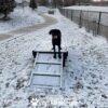 A black dog with an orange ball sits on a snowy Bridge Climb (S/M) 18" Tall, marked by paw prints, beside a fenced park path. Text below says: "PLAY & AGILITY POWERED BY GYMS FOR DOGS.COM.