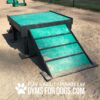 A green and black wooden Bridge Climb (L/X) 24" Tall with ramp sits on gravel by a fence. Text at the bottom reads, "PLAY & AGILITY POWERED BY GYMS FOR DOGS.COM" with a paw print logo.