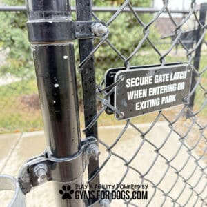 A close-up of a wet chain-link gate in a fenced dog park displays the Double Dog Park Sign Kit—two signs, one reading "Secure gate latch when entering or exiting park"; the other: "Gyms For Dogs.com.