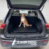 A brown and white dog with a blue collar sits in the open trunk of a black SUV on a Jump Dog SUV Pet Comfort Pad. "JUMP DOG.COM" is visible on the back bumper, ready for their trip to the dog park.