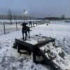 A black dog stands on a Bridge Climb 24" Tall (L/X) wooden platform in a snow-covered, fenced dog park on a winter evening, as streetlights illuminate the snowy landscape and road in the background.