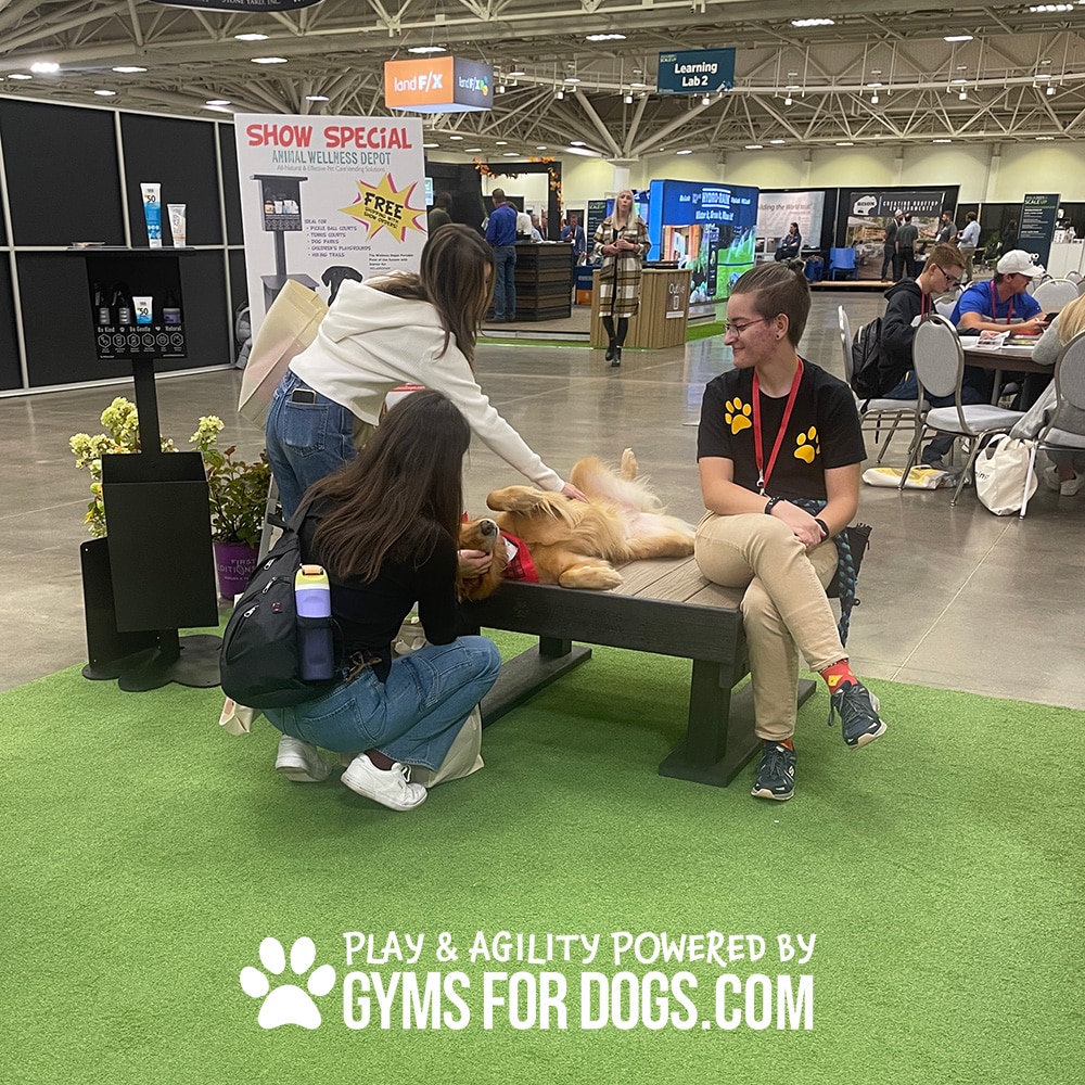 Two women pet a golden retriever lying on its back on a Training Platform (L/X) bench, while another woman in a paw-print shirt smiles nearby. The indoor event features booths, with "PLAY & AGILITY POWERED BY GYMS FOR DOGS.COM" text at the bottom.