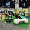 The booth features green turf, a large white bone prop, agility ramps, and two dogs, showcasing the "Training Platform (L/X)." Two staff in blue shirts engage with visitors under a "Healthy Pets" sign, with other booths and attendees in the background.