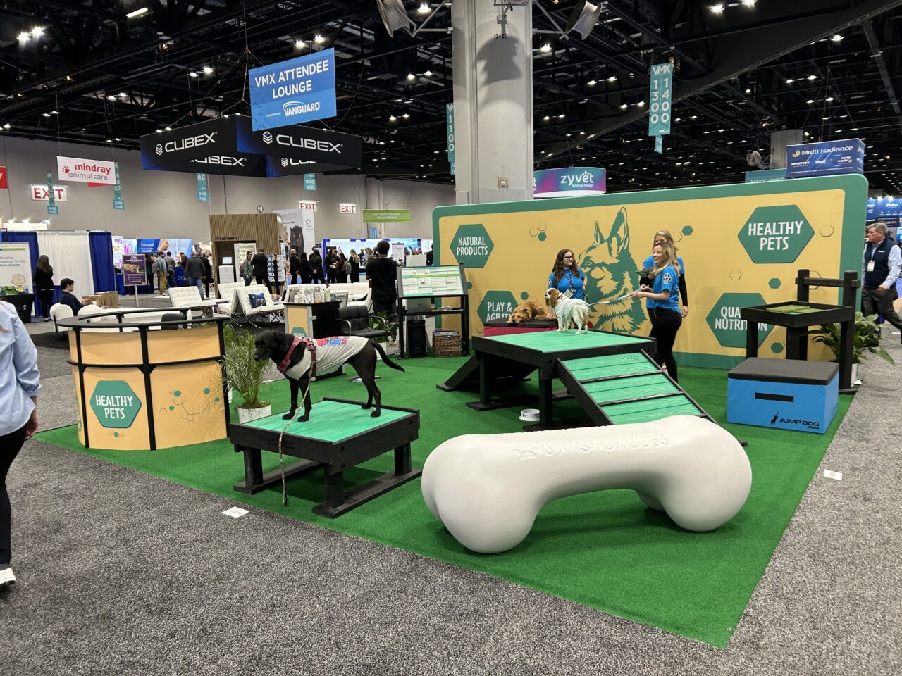 The booth features green turf, a large white bone prop, agility ramps, and two dogs, showcasing the "Training Platform (L/X)." Two staff in blue shirts engage with visitors under a "Healthy Pets" sign, with other booths and attendees in the background.