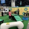 Two women with two dogs stand at a pet expo booth showcasing the Bridge Climb 24" Tall (L/X) agility equipment, which resembles a Dog Park structure. A giant dog bone prop and a "Healthy Pets, Quality Nutrition" backdrop complete the display.