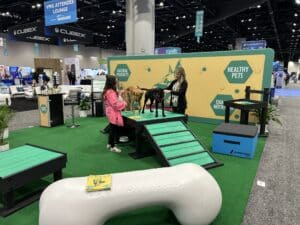 Two women with two dogs stand at a pet expo booth showcasing the Bridge Climb 24" Tall (L/X) agility equipment, which resembles a Dog Park structure. A giant dog bone prop and a "Healthy Pets, Quality Nutrition" backdrop complete the display.