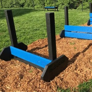 A Jump Hurdle - Small is set up on mulch in a grassy outdoor dog park, likely within a fenced area with green trees in the background.