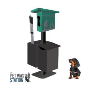 A small black and brown dachshund sits near The Pet Waste Station with Scooper - Surface Mount, a green and black unit featuring a bag dispenser, attached garbage bin, and pet waste scoopers. The logo appears in the bottom left corner.
