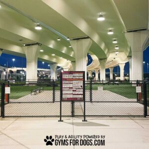 A fenced dog park with green turf sits under a large overpass at dusk, lit by overhead lights. Near the entrance stands the Professional Rules & Wellness Sign - Surface Mount - Vertical Sign. Text at bottom: "Play & Agility Powered by GymsForDogs.com.