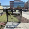 The Service Gate 6' Tall with Self-Closing Hinges & Latch - Doggie Safe Spacing features black metal with dog silhouettes and a dog graphic, welcoming visitors to the Dog Park amid modern buildings, parked cars, and a sidewalk.