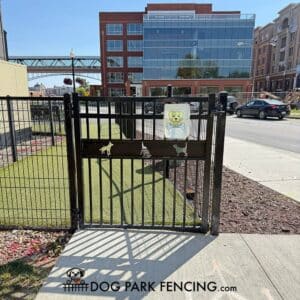 The Service Gate 6' Tall with Self-Closing Hinges & Latch - Doggie Safe Spacing features black metal with dog silhouettes and a dog graphic, welcoming visitors to the Dog Park amid modern buildings, parked cars, and a sidewalk.