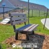 A Trail Bench w/ Back - 4' (Surface Mount) sits on mulch near a sidewalk, with a black fence and agility ramp behind. It's a sunny day with clear blue sky. Text: "Play & agility powered by GYMSFORDOGS.COM.
