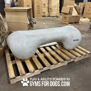 A large, antique polar gray Agility Bone (L/X) by GymsForDogs.com sits on a pallet in a warehouse with boxes and straw bales. Text reads: "Play & Agility Powered by GymsForDogs.com.