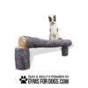 A black and white dog sits on a large, realistic fake log bench. Text below reads "Play & Agility Powered by GymsForDogs.com" with paw print icons.