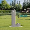 A Cool Dog Water Fountain - STE - Dog Basin stands on a grassy field in a park, with trees and a playground behind it. The fountain features two accessible heights, and the "Cool Dog Water Fountains" logo is visible.