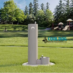 A Cool Dog Water Fountain - STE - Dog Basin stands on a grassy field in a park, with trees and a playground behind it. The fountain features two accessible heights, and the "Cool Dog Water Fountains" logo is visible.