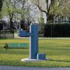 The Cool Dog Water Fountain - STE - Dog Basin & ADA Basin is a blue outdoor fountain with adult and dog-height spouts, set on grass in a park with trees and a black fence. "Cool Dog Water Fountains.com" appears on the image.