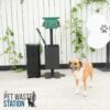 A small brown and white dog stands on pavement near a pet waste station, which includes a green waste bag dispenser and a black trash bin. A white fence and leafy plants are in the background.
