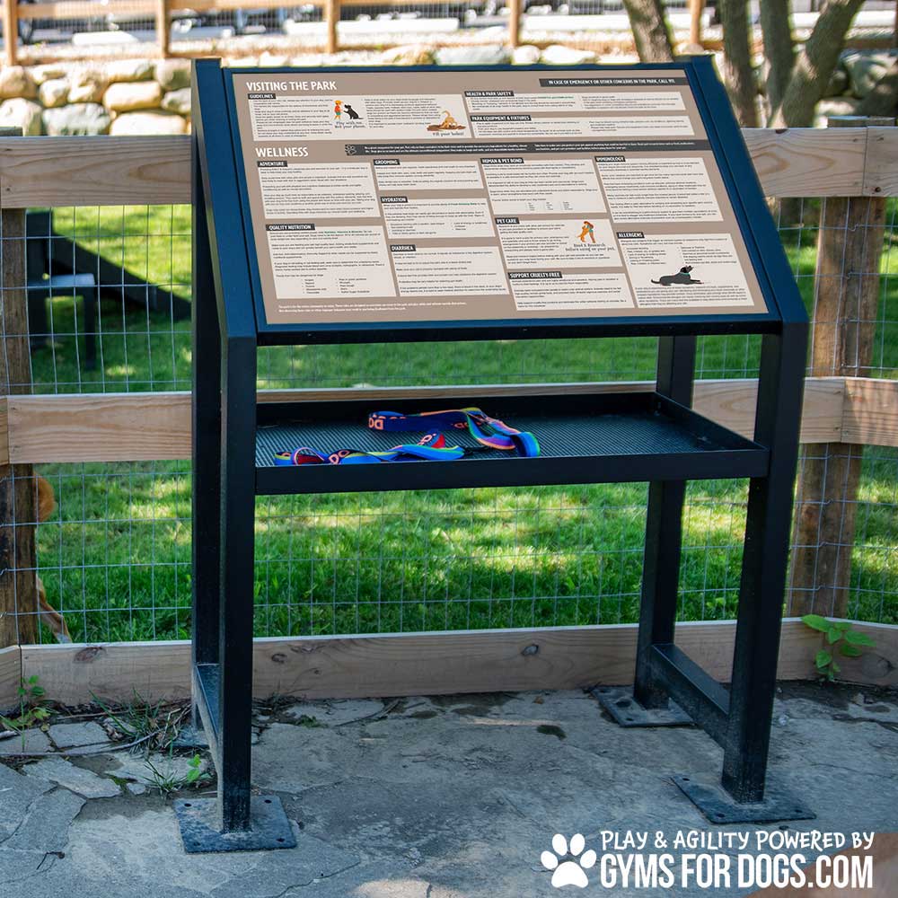 An informational sign with guidelines and illustrations for visiting a dog park sits on a metal stand above a shelf holding a leash and collar. The sign reads “Gyms for Dogs” at the bottom right corner.