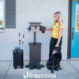 A woman in a yellow shirt stands beside her black dog and The Pet Waste Station - Free Standing, pulling a waste bag from the dispenser near a black trash bin with tools, all positioned by a light brick wall.