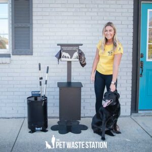 A woman in a yellow shirt smiles while petting a black dog next to The Scooper Station—Storage Box with Scooper for Pet Waste Station—in front of a gray brick building with a blue door.