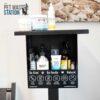 A black pet waste station holds various pet care products, including sprays, creams, and a bottle, with "Be Kind," "Be Gentle," and "Natural" labels. "The Pet Waste Station" logo is visible on the wall behind.