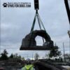A large artificial stone arch is being lifted by a crane at an outdoor construction site. A worker in a safety vest stands below, looking up. The sky is overcast and trees are in the background. "Gyms for Dogs" logo is at the top.