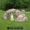 A black and white dog sits under the Motion Mountain Climb - Interactive Play Series in a grassy park with trees and white benches. Text at the bottom reads, "Play & agility powered by gymsfordogs.com.