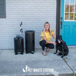 A woman in a yellow shirt kneels by a black dog outside a building with a blue door. Next to them are two Park Waste Caddy small waste receptacles with sanitary liners and decorative lids, plus a waste bag dispenser.