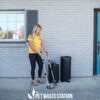 A woman in a yellow shirt uses a scooper to clean pet waste outside a building, standing next to two black waste bins labeled "Pet Waste Station" on a concrete sidewalk.